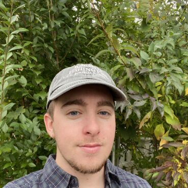 Photo of Cameron wearing a light green baseball cap. He's standing in front of a bushy plant.